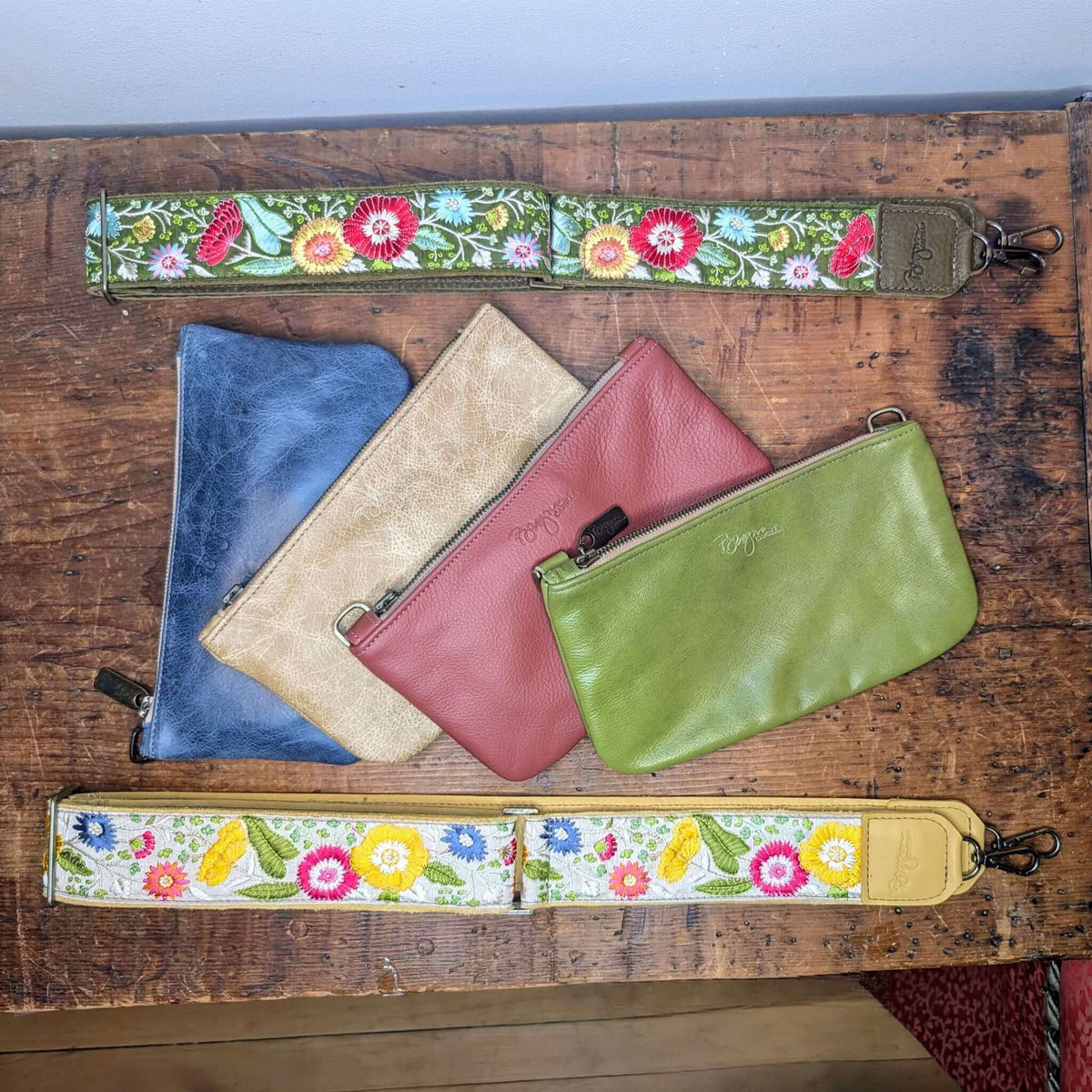 Colorful leather pouches with a floral guitar purse strap on a wooden surface, showcasing the Lizzy Guitar Bag Strap - Evergreen Wildflowers.